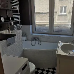 Jasna łazienka w stylu retro z wanną obudowaną mozaiką, umywalką na szafce i pralką, czarno-biała podłoga w szachownicę, widok z okna na budynek.