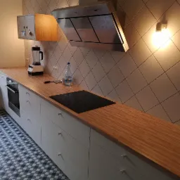 Nowoczesna kuchnia z drewnianym blatem, białymi szafkami, indukcyjną płytą grzewczą i geometryczną podłogą w odcieniach szarości i błękitu, widziana pod kątem.