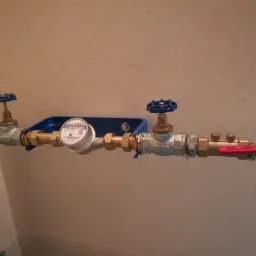 Zamontowany wodomierz z niebieskimi zaworami i przyłączami na ścianie o jasnym odcieniu beżu.