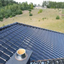 Błyszczący, czarny dach z dachówki ceramicznej z widoczną instalacją odgromową i kominem wentylacyjnym, na tle pagórkowatego terenu z budynkiem gospodarczym.