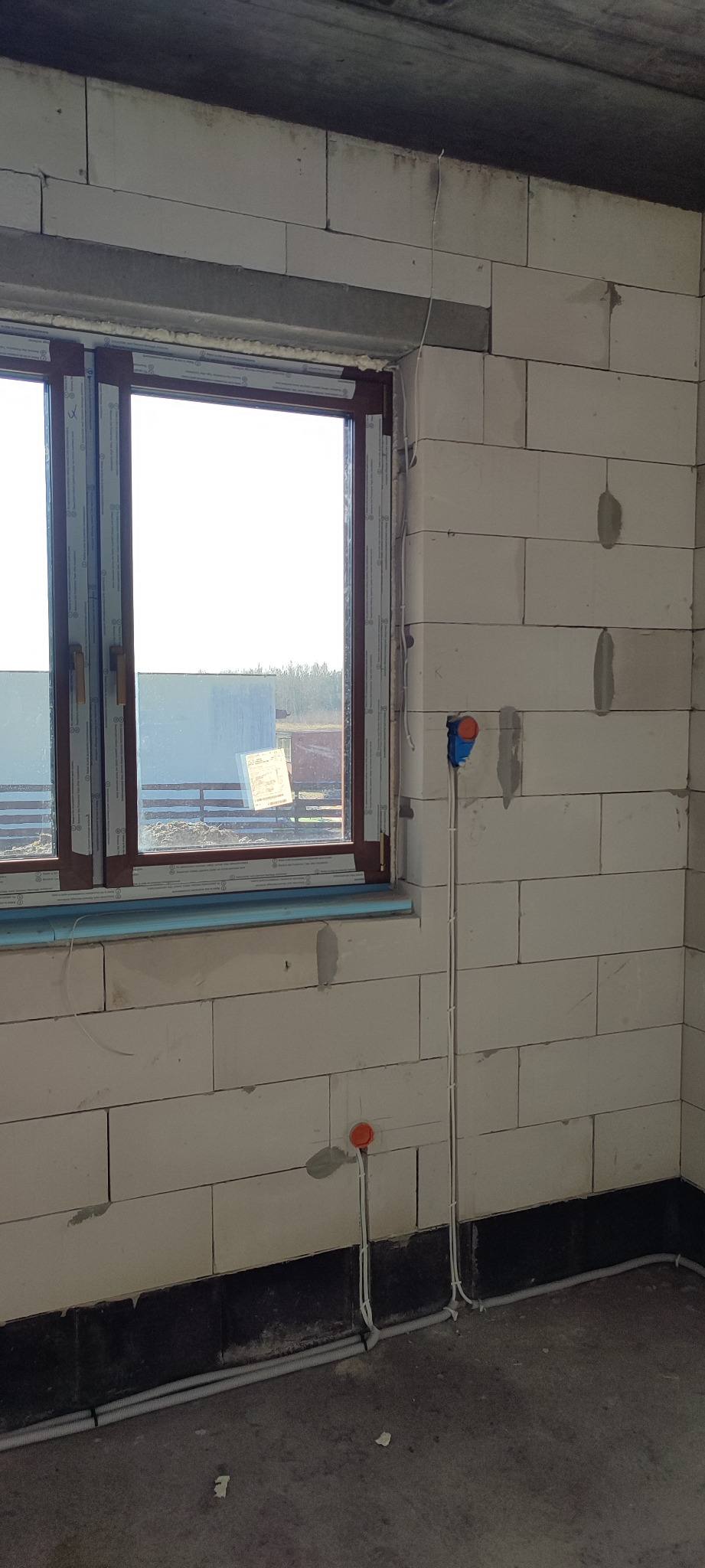 Surowy stan wnętrza z widocznymi przewodami elektrycznymi, puszkami instalacyjnymi w kolorze pomarańczowym i niebieskim, oraz rurami osłonowymi przy podłodze, ściany z białych bloczków.