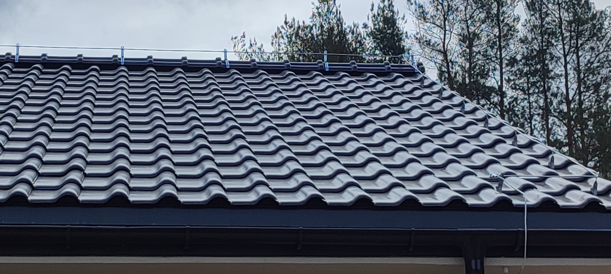 Nowy, grafitowy dach z dachówki ceramicznej z zamontowanym systemem odgromowym, widok z dołu na tle drzew.