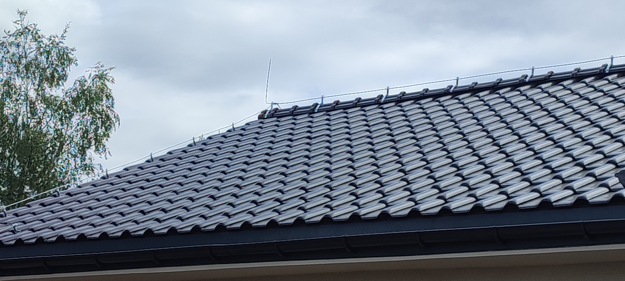 Ciemnoszara dachówka ceramiczna z zamontowaną instalacją odgromową, widok z dołu na tle pochmurnego nieba i fragmentu drzewa.