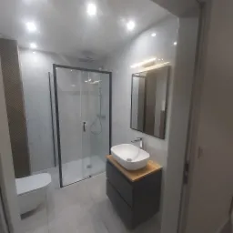 Nowoczesna łazienka po remoncie z kabiną prysznicową typu walk-in z czarnymi ramami, umywalką nablatową na szafce z drewnianym blatem, i toaletą podwieszaną.