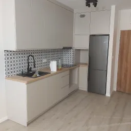 Jasna kuchnia z drewnianym blatem, czarną armaturą i wzorzystą mozaiką nad blatem, uzupełniona o lodówkę i drewniane drzwi w tle.