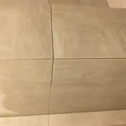 Beżowe płytki podłogowe ułożone w regularny wzór z wyraźnymi fugami, widoczne drobne zabrudzenia na powierzchni płytek.