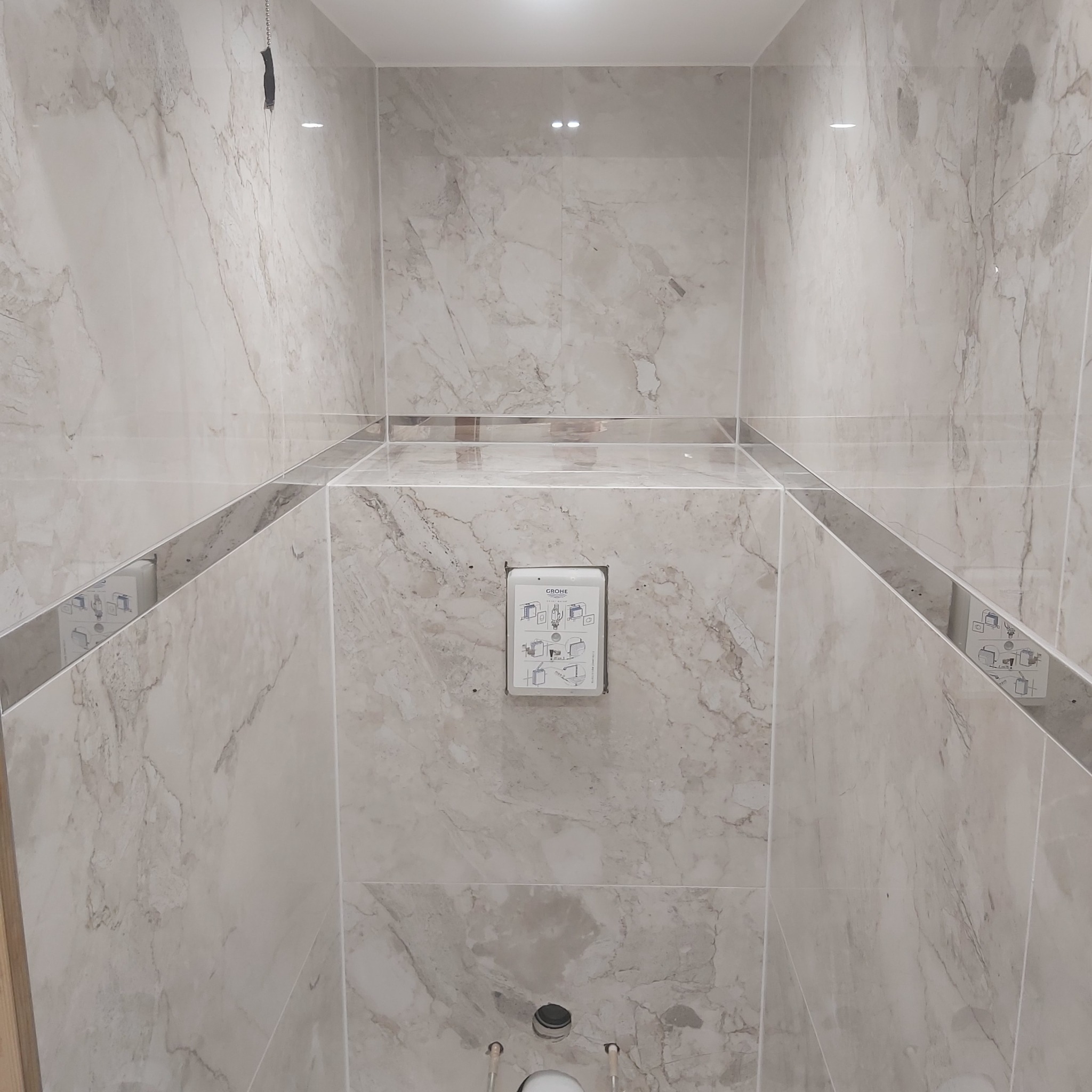 Wnęka w łazience wyłożona płytkami imitującymi marmur, z widoczną podtynkową spłuczką Grohe i przygotowanym odpływem toalety.