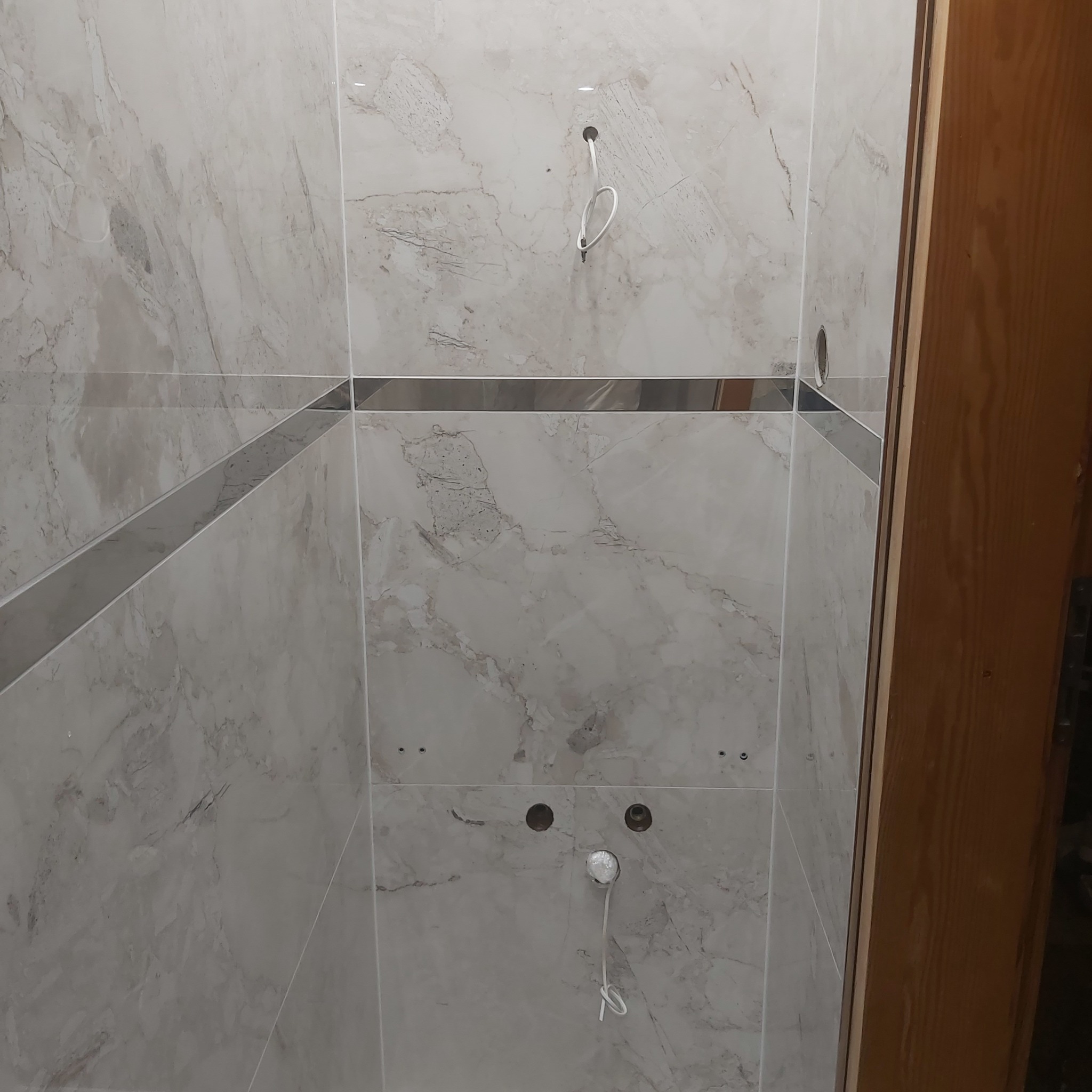 Ściana łazienki wyłożona marmurkowymi płytkami z metalowymi listwami, widoczne otwory na armaturę i wystające przewody.