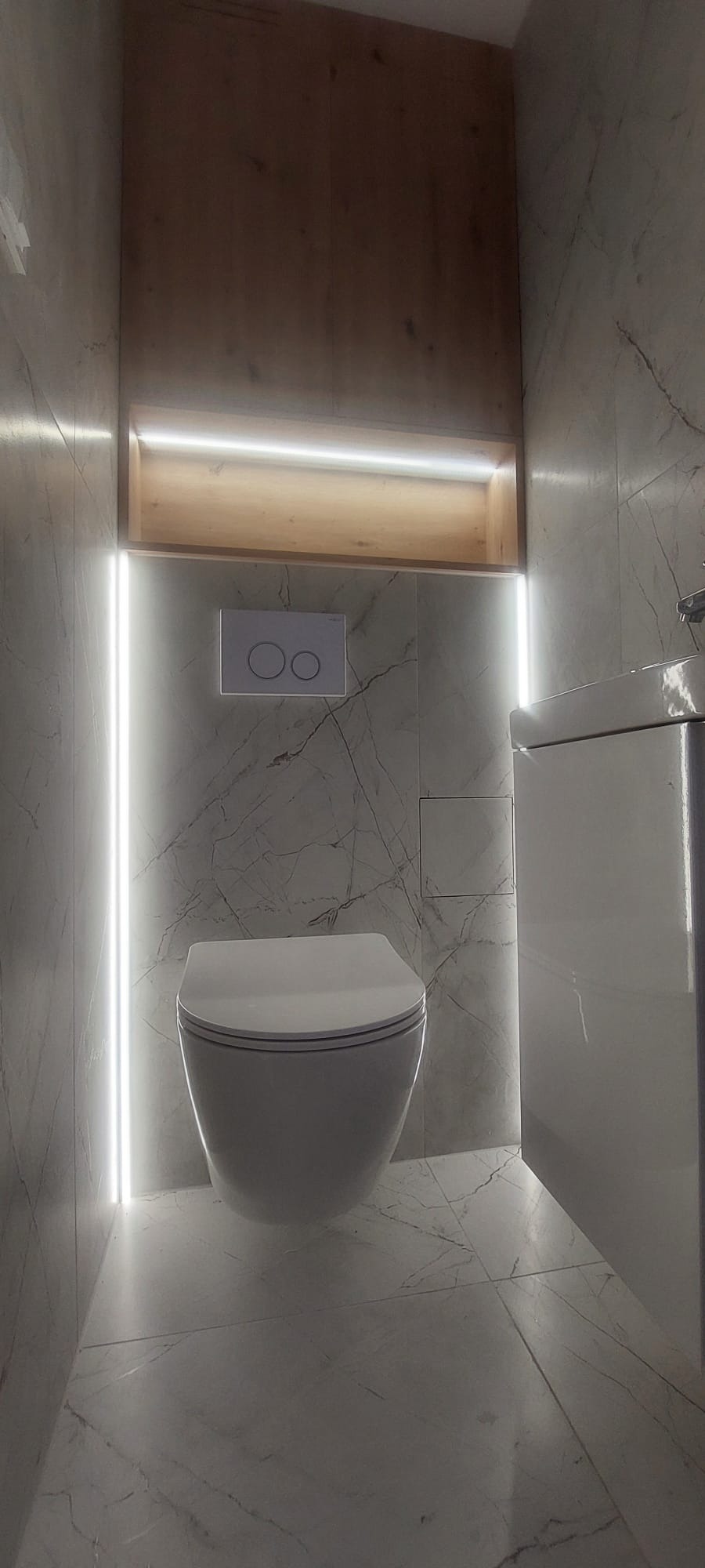 Nowoczesna toaleta podwieszana w łazience wykończonej marmurowymi płytkami, z podświetleniem LED wzdłuż pionowych krawędzi i górnej wnęki z drewnianą półką.
