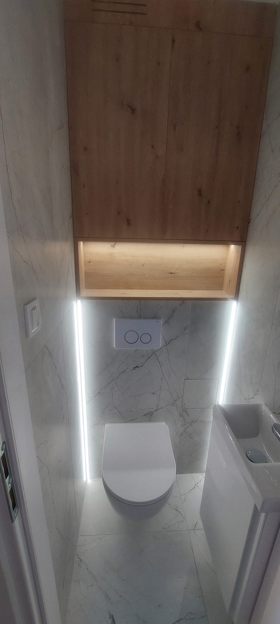 Nowoczesna, mała łazienka z marmurowymi płytkami, podświetleniem LED, wiszącą toaletą i szafką z drewnianymi frontami nad nią.