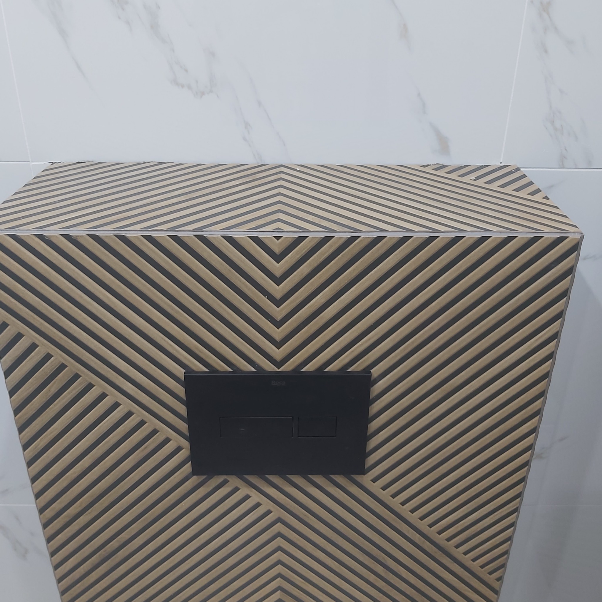 Kompaktowa zabudowa stelaża WC wykończona płytkami z geometrycznym wzorem w odcieniach beżu i szarości, na tle białych płytek imitujących marmur, z widocznym czarnym panelem sterującym Roca.
