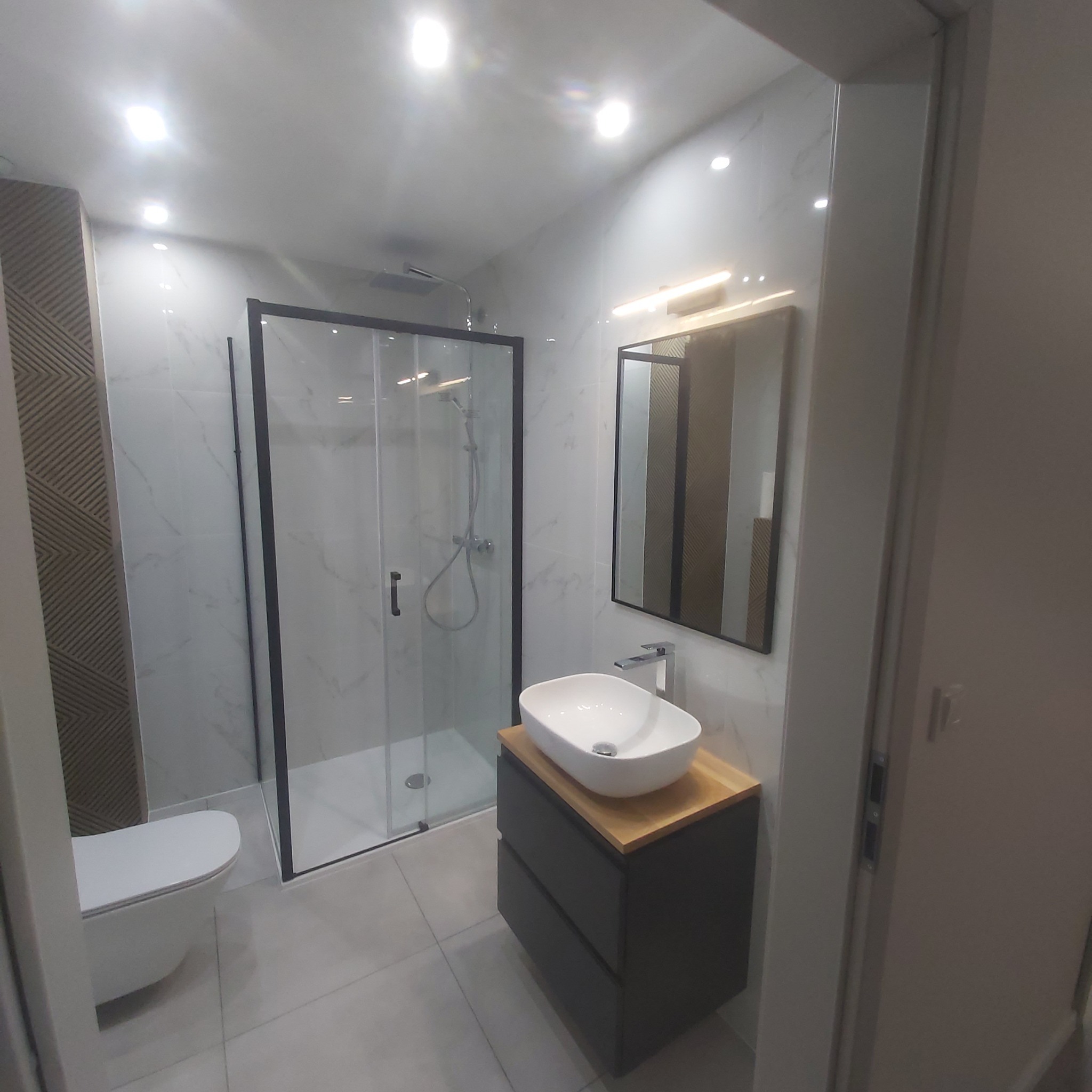 Nowoczesna łazienka po remoncie z kabiną prysznicową typu walk-in z czarnymi ramami, umywalką nablatową na szafce z drewnianym blatem, i toaletą podwieszaną.