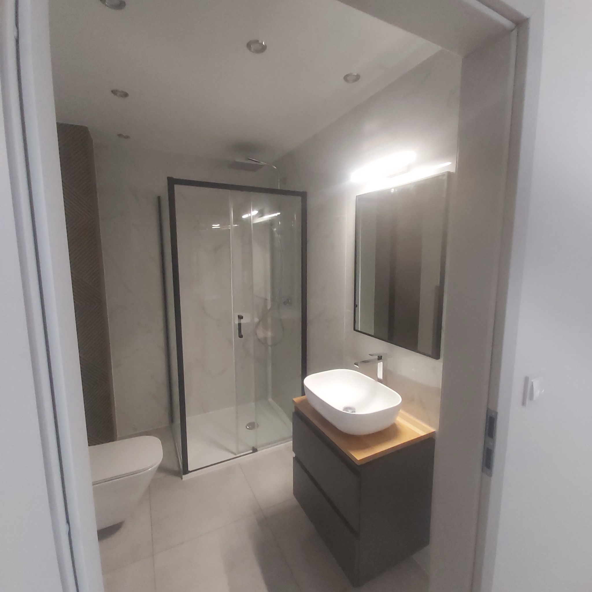 Nowoczesna łazienka z kabiną prysznicową w czarnej ramie, umywalką nablatową na szafce z szufladami i podświetlanym lustrem, utrzymana w jasnej kolorystyce.