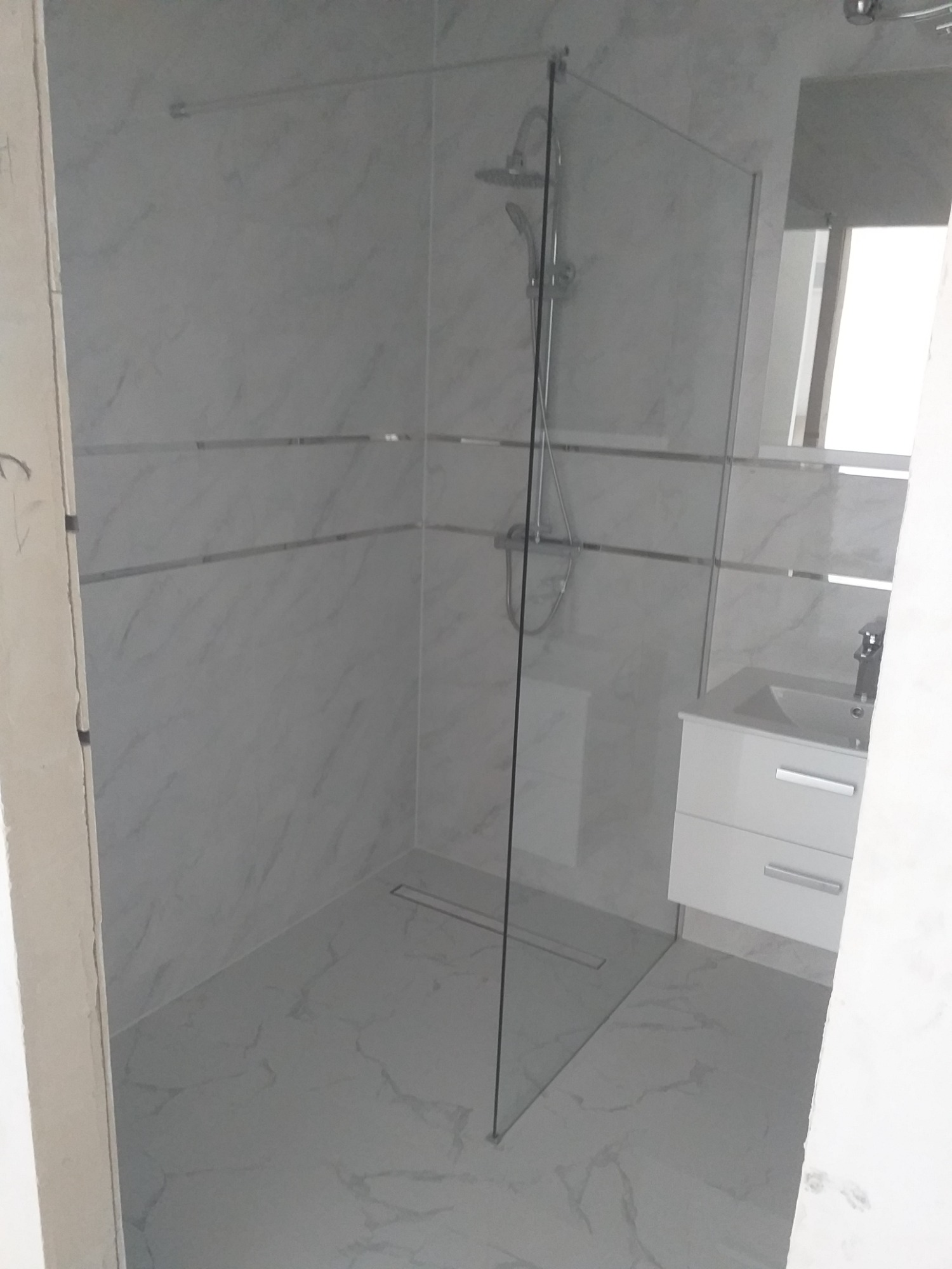 Nowoczesna łazienka z kabiną prysznicową typu walk-in, wykończona białymi płytkami imitującymi marmur, z widocznym odpływem liniowym w podłodze i białą szafką pod umywalką z dwiema szufladami.