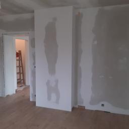 Usługi Ogólnobudowlane Robert Kolka - Wnętrze w trakcie wykańczania: ściany po szpachlowaniu, widoczne łączenia płyt karton-gips, drewniana podłoga, drabina w przejściu do innego pomieszczenia.