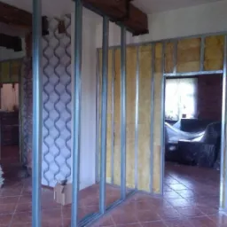 Montaż metalowego stelażu ścian działowych z izolacją z wełny mineralnej w trakcie prac remontowych, widoczne płyty kartonowo-gipsowe i dekoracyjna struktura na fragmencie ściany.