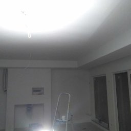 Usługi Ogólnobudowlane Robert Kolka - Wnętrze w trakcie prac wykończeniowych: świeżo pomalowany sufit i ściany na biało, widoczna drabina malarska z kubkiem na górze, wystające kable ze ściany, okna przesuwne.