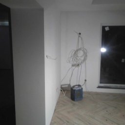 Usługi Ogólnobudowlane Robert Kolka - Wnętrze w trakcie remontu: świeżo pomalowane ściany na biało, widoczne kable elektryczne zwisające ze ściany, podłoga parkietowa w jodełkę, okno z widocznym światłem zewnętrznym oraz niebieski...