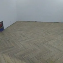 Ułożona w jodełkę podłoga z płytek imitujących drewno w pustym, jasnym pomieszczeniu z widoczną instalacją elektryczną w ścianie.