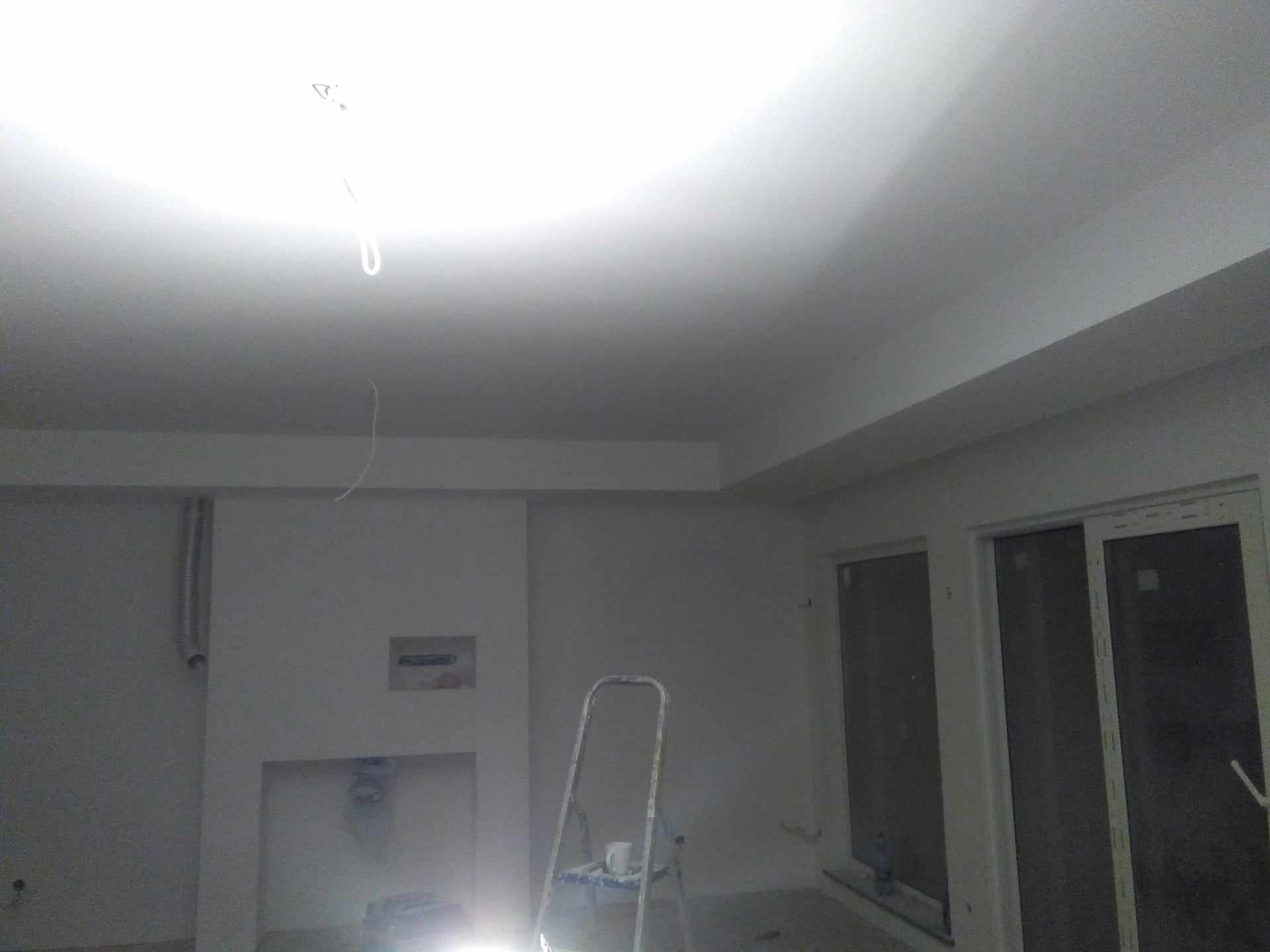 Wnętrze w trakcie prac wykończeniowych: świeżo pomalowany sufit i ściany na biało, widoczna drabina malarska z kubkiem na górze, wystające kable ze ściany, okna przesuwne.