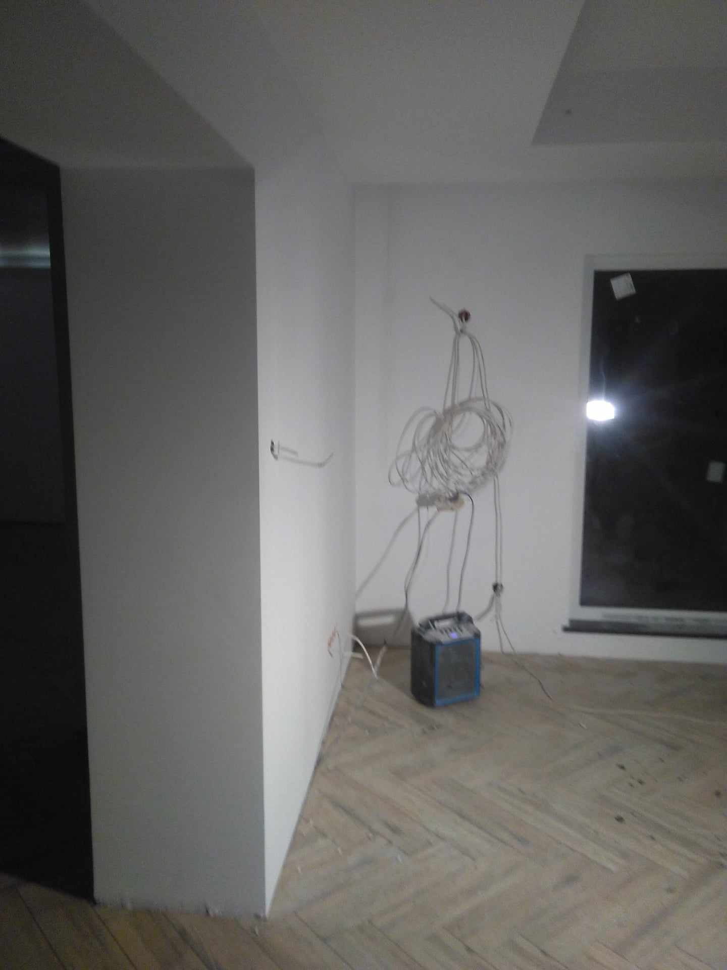 Wnętrze w trakcie remontu: świeżo pomalowane ściany na biało, widoczne kable elektryczne zwisające ze ściany, podłoga parkietowa w jodełkę, okno z widocznym światłem zewnętrznym oraz niebieski...