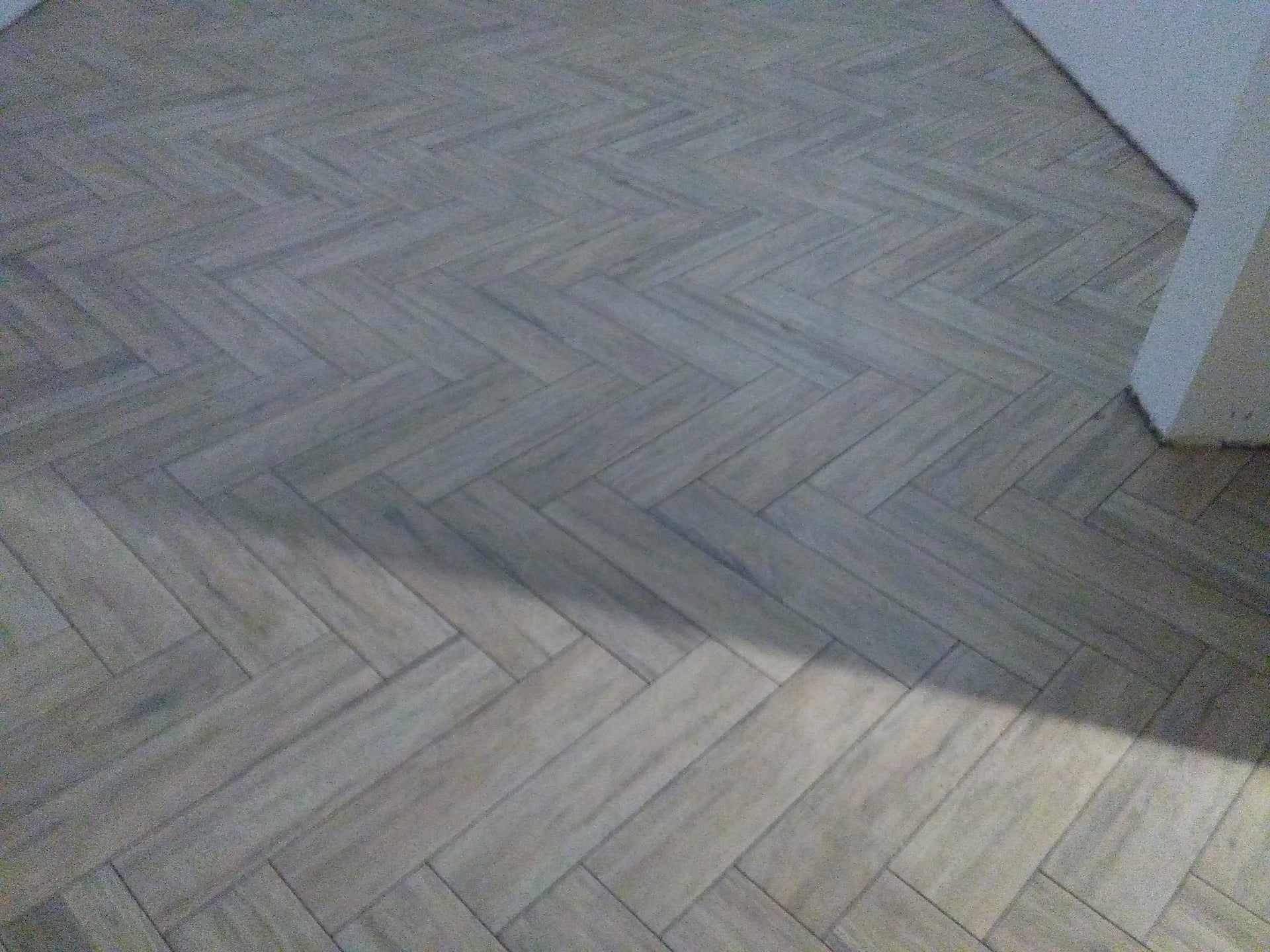 Ułożona w jodełkę podłoga z płytek imitujących drewno w odcieniach szarości, widoczny fragment ściany w rogu pomieszczenia.