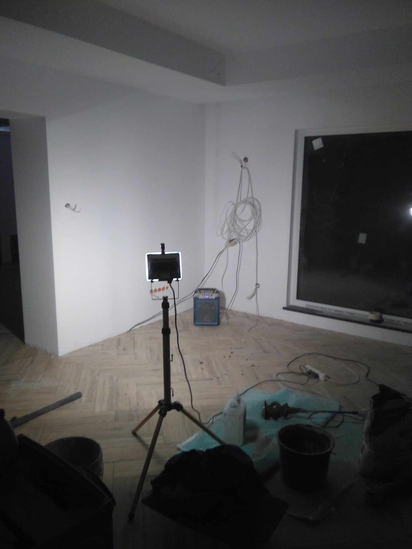 Wnętrze w trakcie prac wykończeniowych, widoczne narzędzia, kabel zwisający ze ściany, lampa budowlana na statywie i częściowo ułożona podłoga z paneli imitujących drewno.