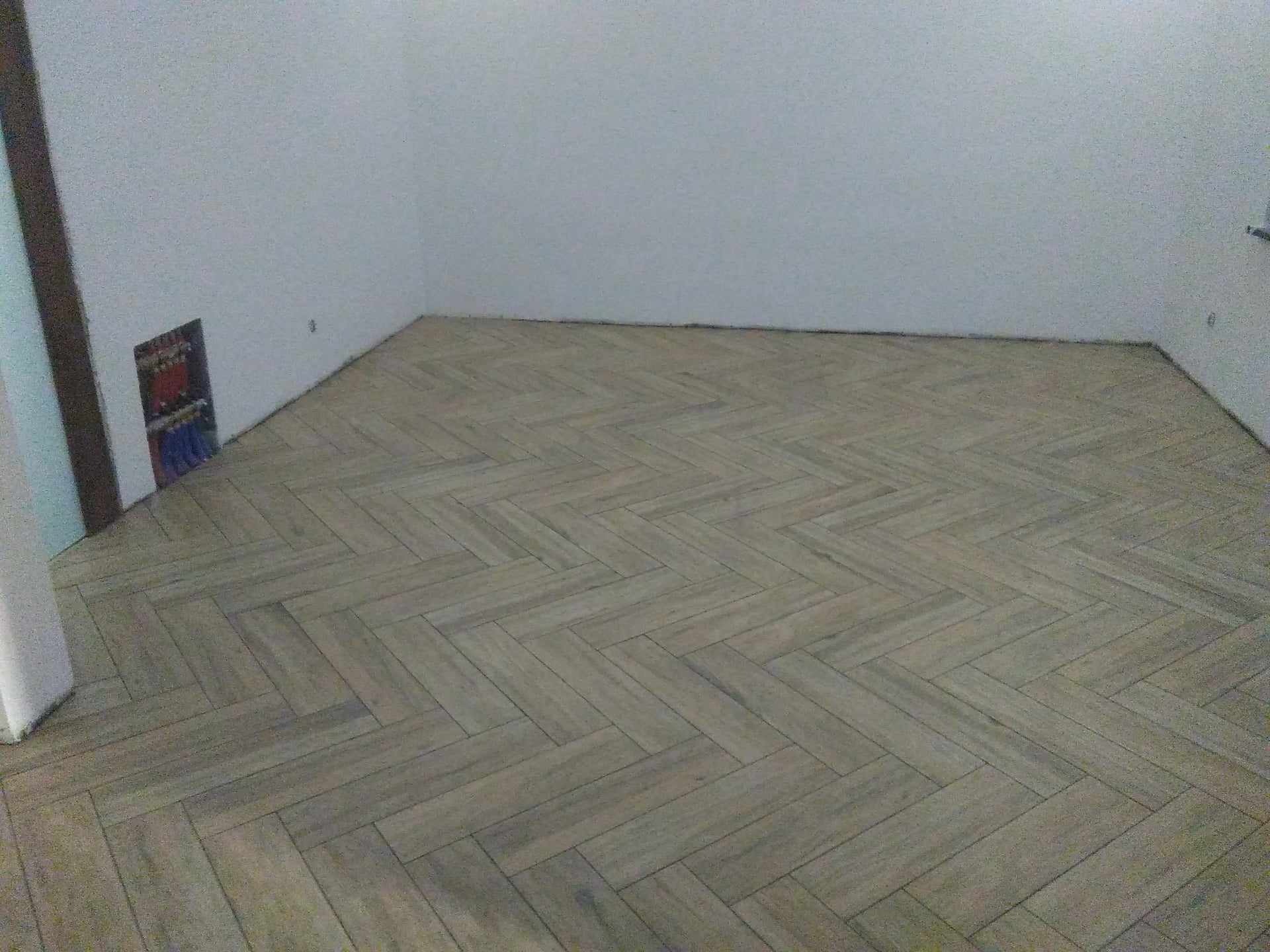 Ułożona w jodełkę podłoga z płytek imitujących drewno w pustym, jasnym pomieszczeniu z widoczną instalacją elektryczną w ścianie.