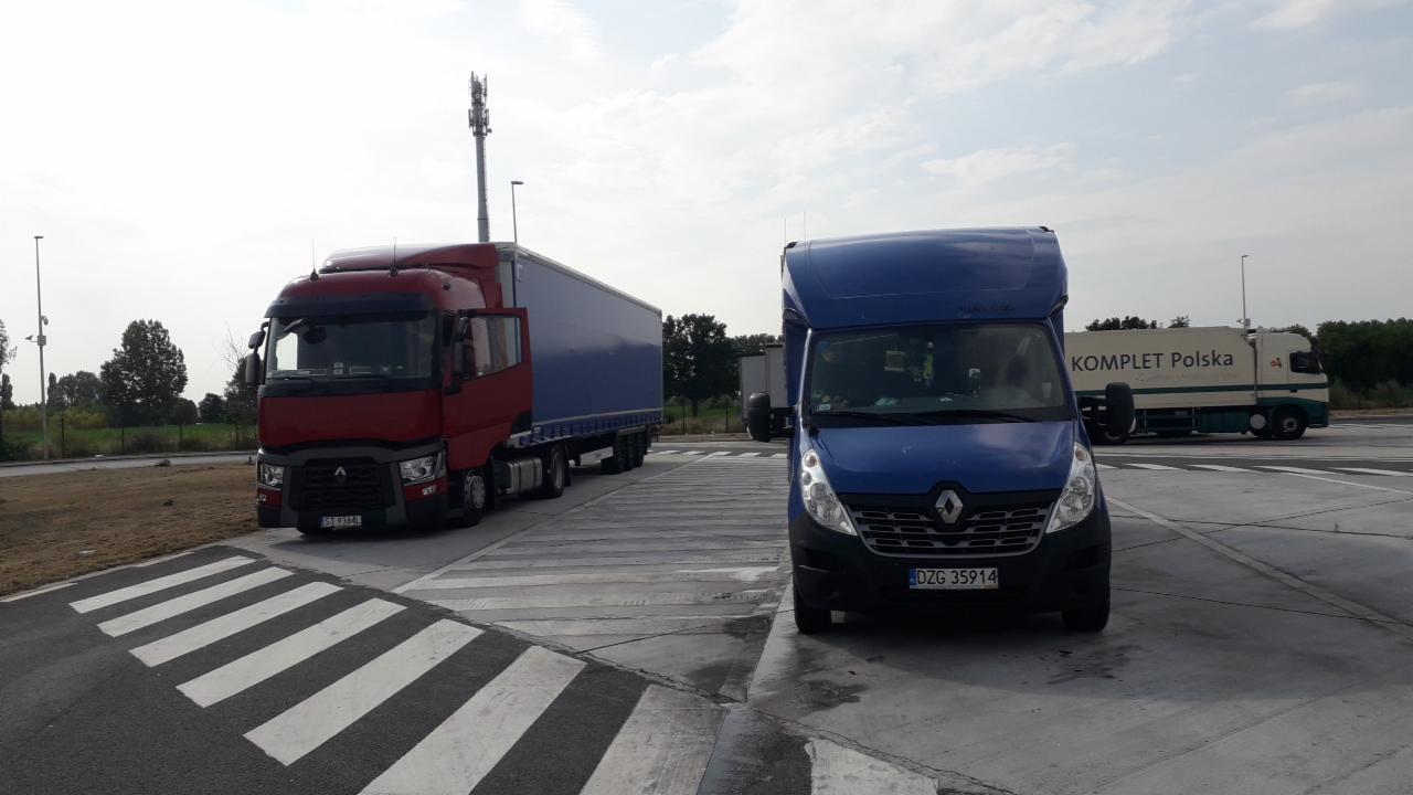 Czerwona ciężarówka z niebieską naczepą i niebieski bus Renault Master zaparkowane obok siebie na parkingu dla tirów, w tle inne pojazdy i maszt telefonii komórkowej.