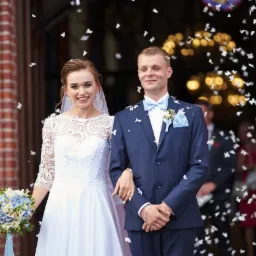 Uśmiechnięta para młoda wychodzi z kościoła podczas sypania konfetti, panna młoda w białej sukni z koronkową górą, pan młody w granatowym garniturze z błękitną muchą i butonierką.