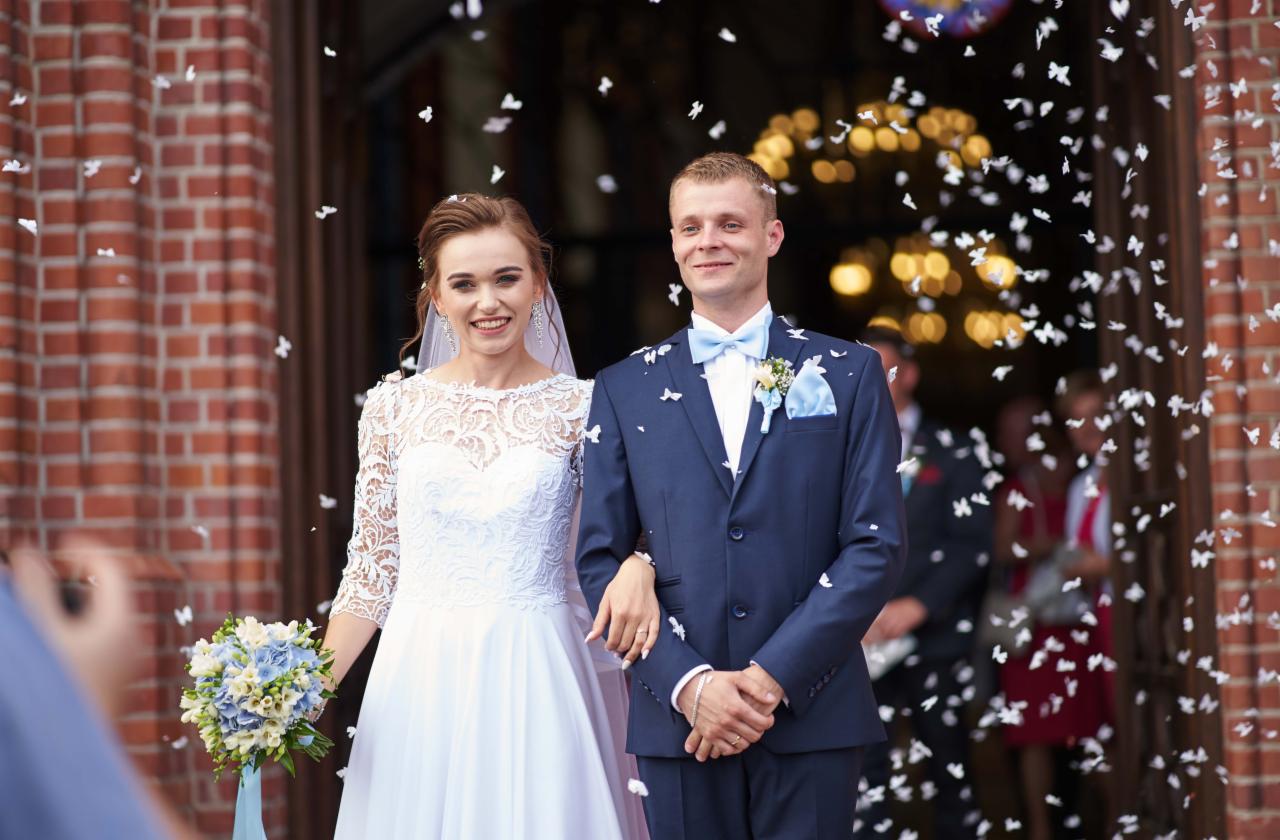 Uśmiechnięta para młoda wychodzi z kościoła podczas sypania konfetti, panna młoda w białej sukni z koronkową górą, pan młody w granatowym garniturze z błękitną muchą i butonierką.