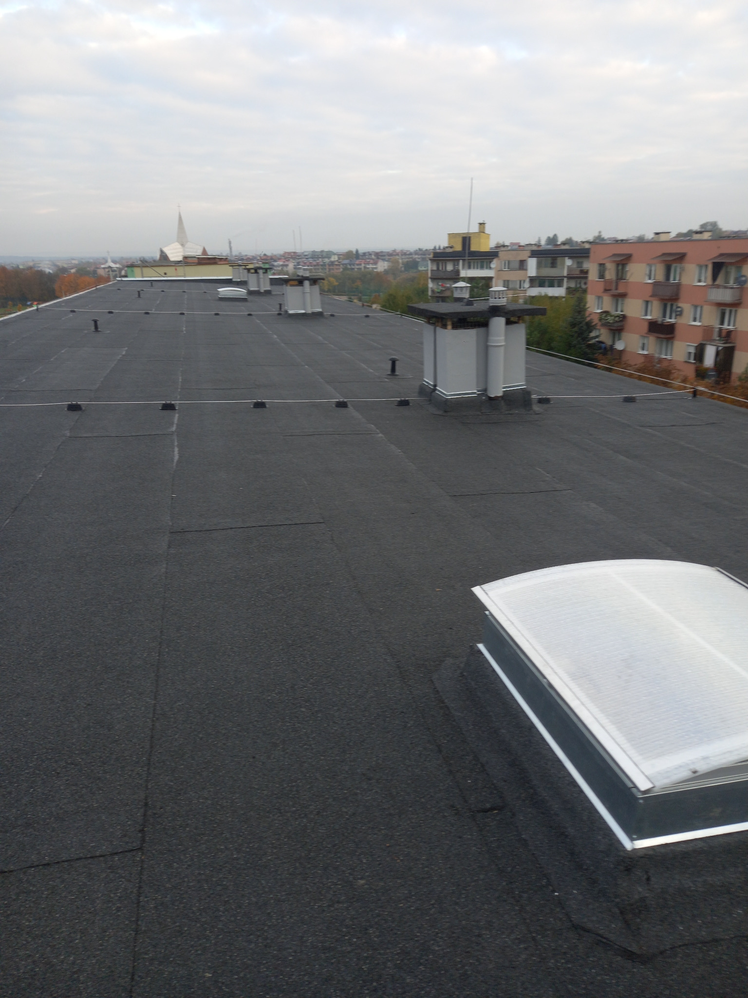 Wykończony dach płaski z papy termozgrzewalnej z świetlikiem dachowym i kominkami wentylacyjnymi na tle zabudowy miejskiej w pochmurny dzień.