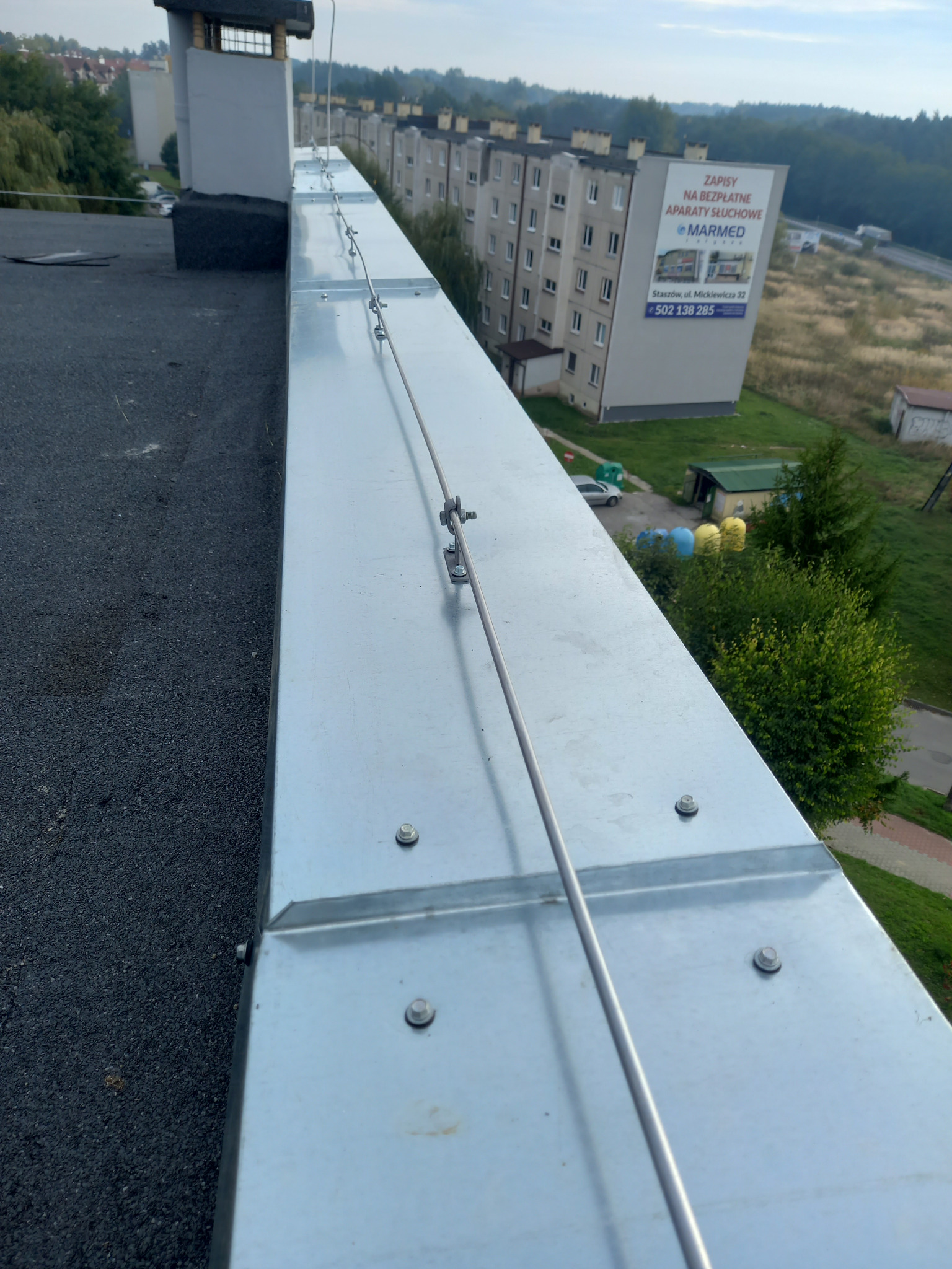 Metalowa instalacja odgromowa na dachu budynku, zabezpieczona śrubami. W tle widoczny blok mieszkalny i zieleń. Ujęcie z góry, podkreślające detale montażu.