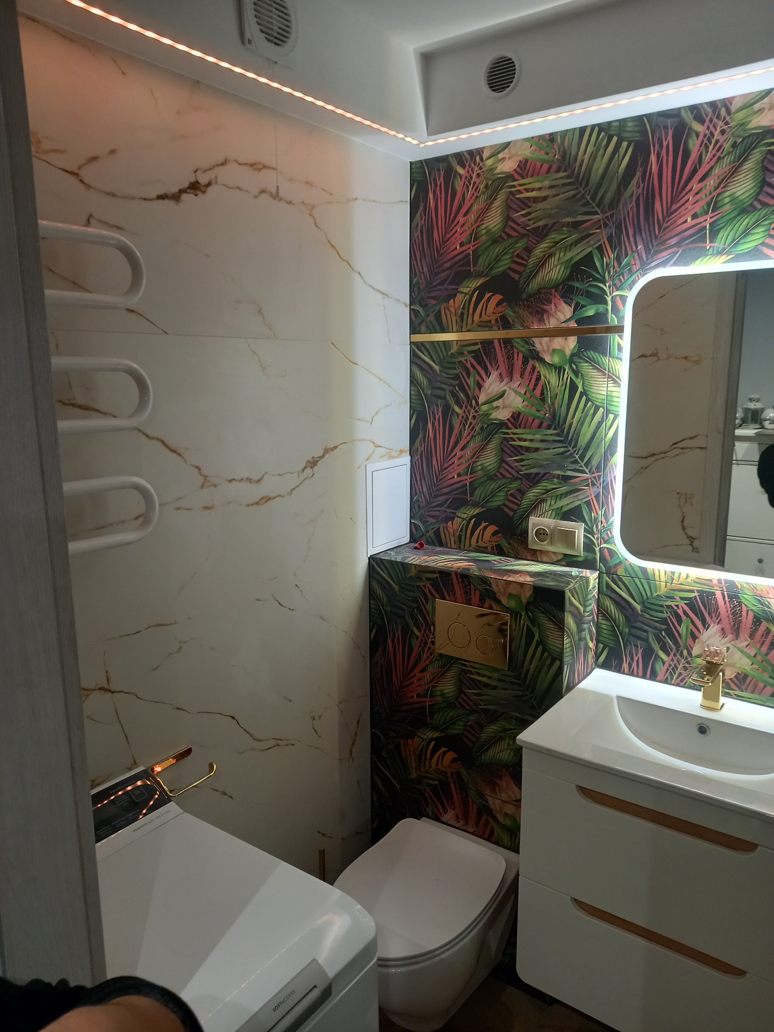 Nowoczesna łazienka z marmurowymi płytkami i tropikalnym motywem na ścianie, podświetlana listwą LED. Uwagę przykuwa złota armatura i detale.