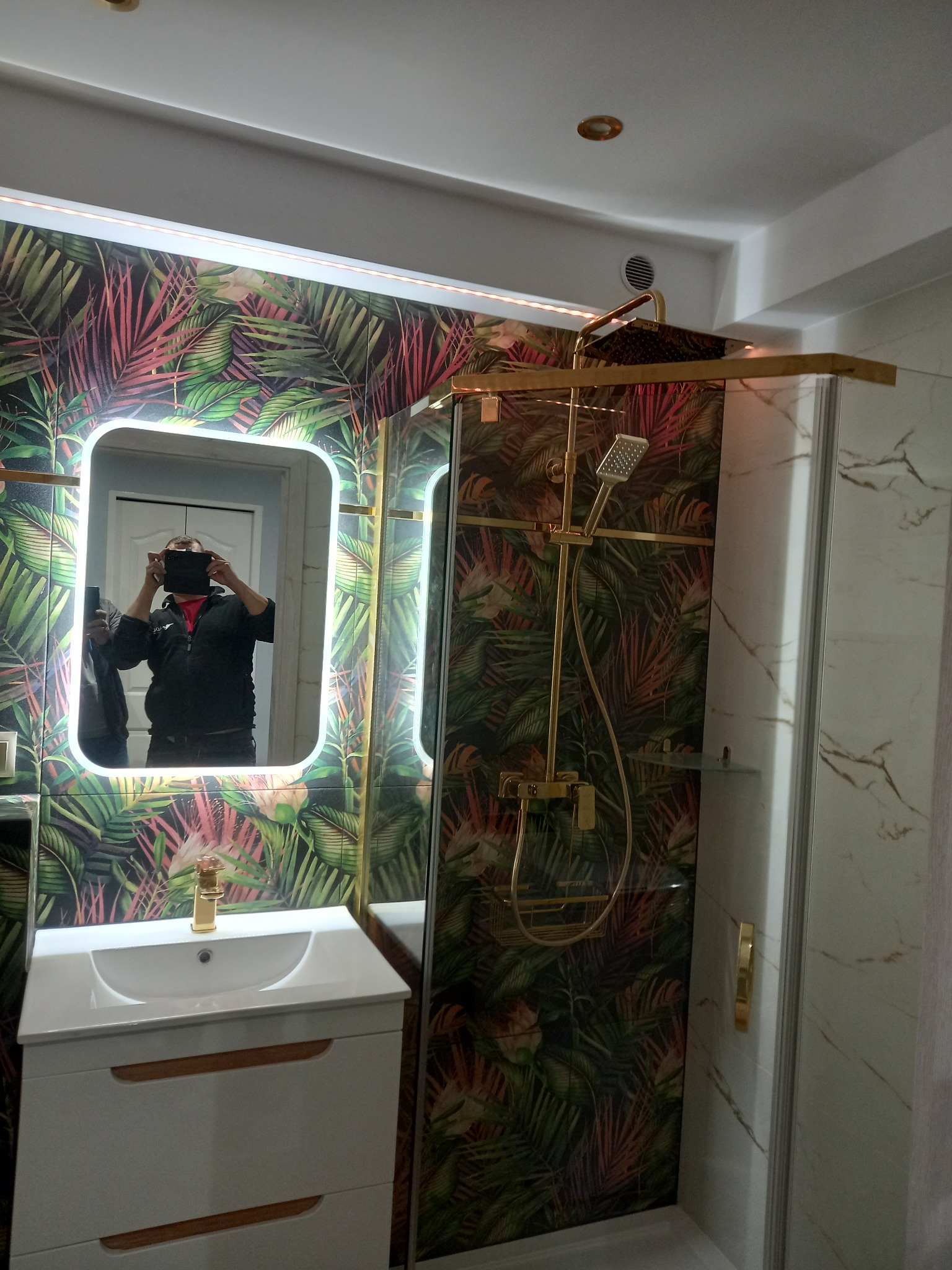 Wykończona łazienka z kabiną prysznicową i umywalką w szafce. Na ścianach tapeta w tropikalne liście, złota armatura i oświetlenie LED.