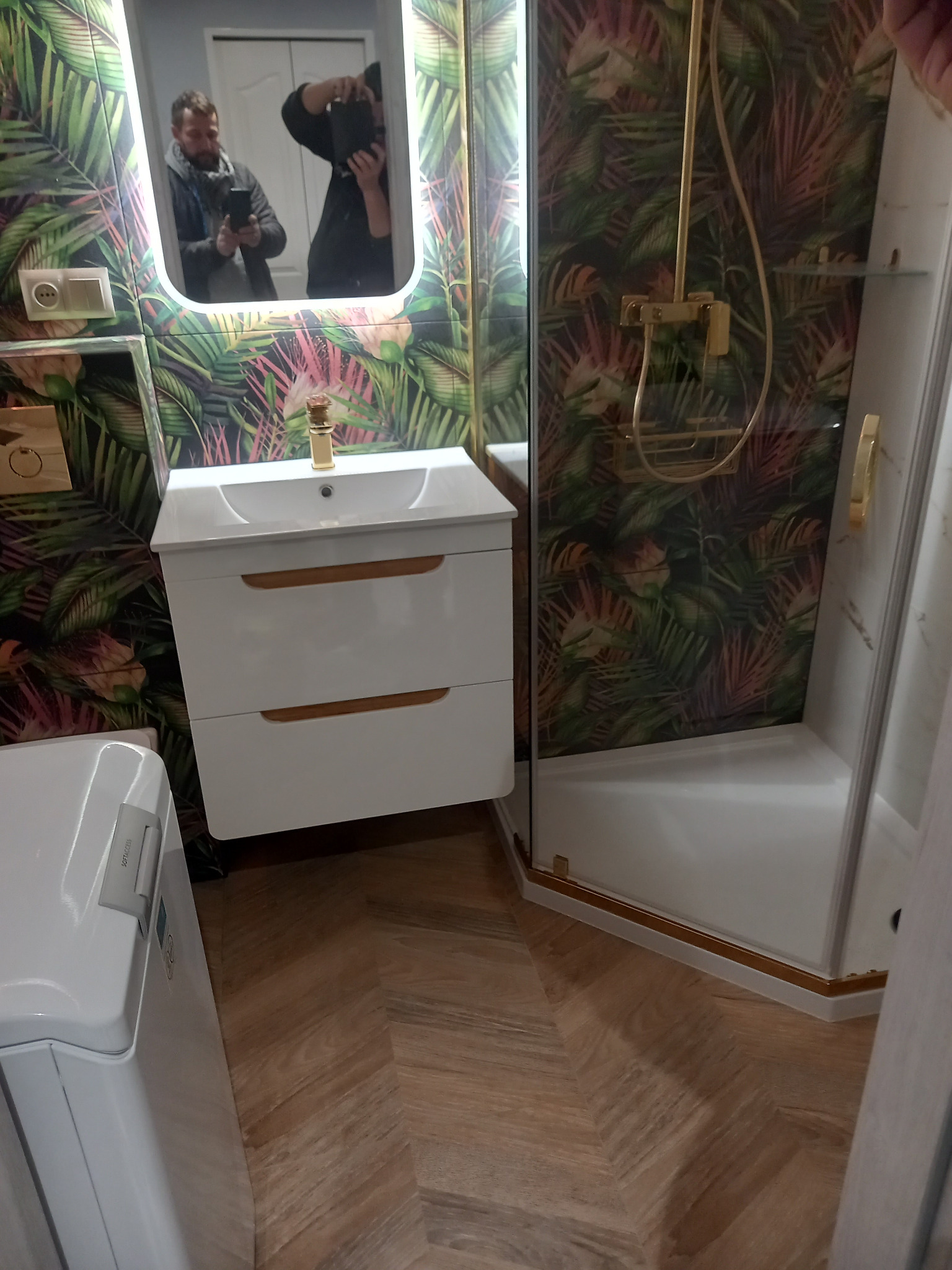 Nowoczesna łazienka po remoncie: biała szafka z umywalką, kabina prysznicowa ze złotymi akcentami, wzorzyste tapety w tropikalne liście i podłoga imitująca drewno.