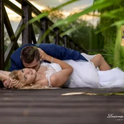 Panna młoda w koronkowej sukni leży na drewnianym pomoście, oblubieniec w granatowym garniturze całuje ją w szyję, w tle trzciny i zachodzące słońce, widoczna nagroda Orły Branży Ślubnej 2019.