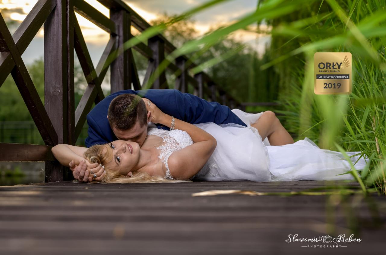 Panna młoda w koronkowej sukni leży na drewnianym pomoście, oblubieniec w granatowym garniturze całuje ją w szyję, w tle trzciny i zachodzące słońce, widoczna nagroda Orły Branży Ślubnej 2019.