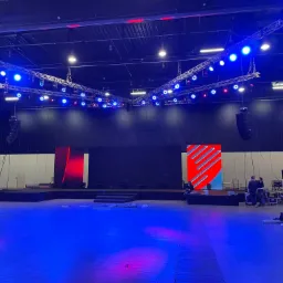 Przygotowania do wydarzenia w Warszawie: montaż oświetlenia scenicznego z dominującym niebieskim światłem, widoczna konstrukcja kratownicowa pod sufitem oraz ekrany LED w kolorach czerwonym...