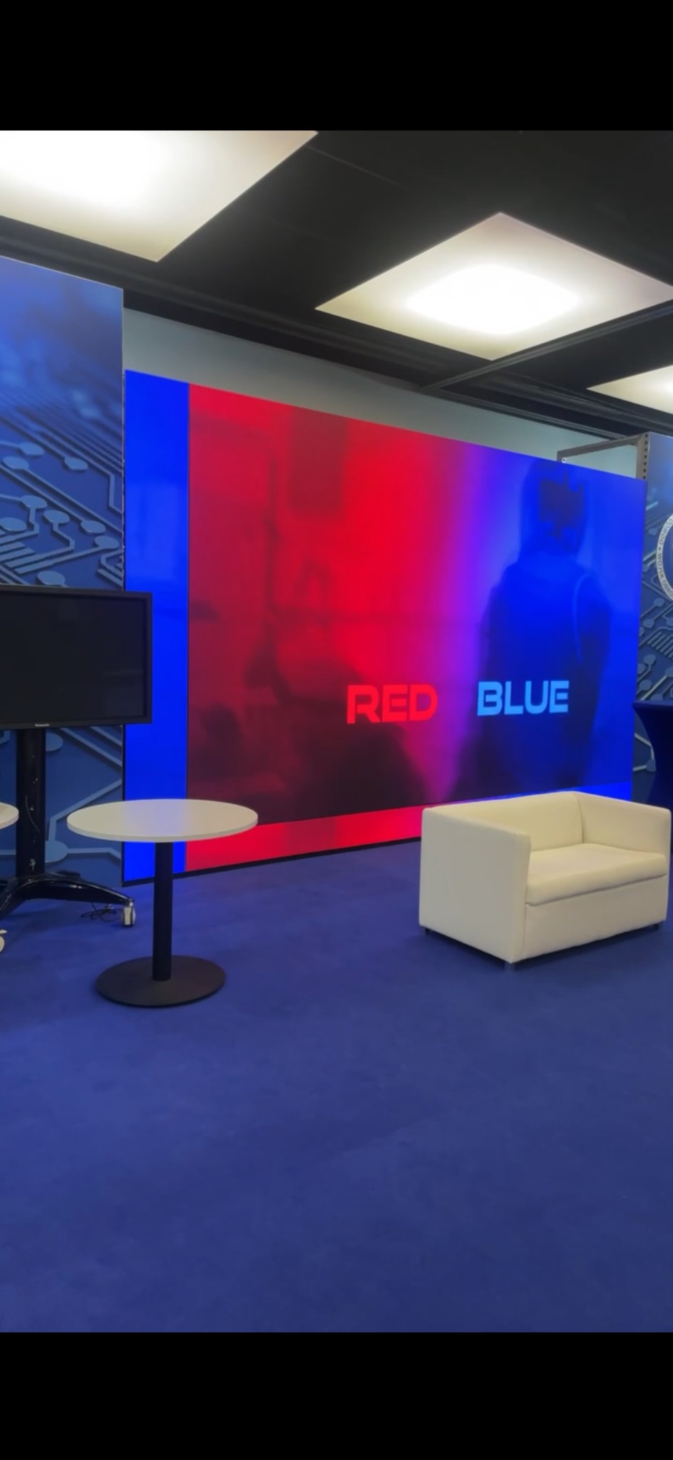 Aranżacja przestrzeni eventowej z dużym ekranem LED wyświetlającym napisy 'RED' i 'BLUE', białą sofą i stolikami na niebieskiej wykładzinie.