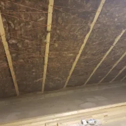 Ocieplenie poddasza wełną mineralną z drewnianą konstrukcją nośną i rozwieszoną pomarańczową nicią mocującą, widok od dołu na surowy betonowy strop.