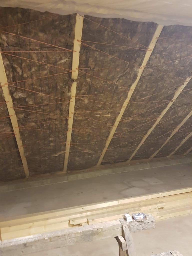 Ocieplenie poddasza wełną mineralną z drewnianą konstrukcją nośną i rozwieszoną pomarańczową nicią mocującą, widok od dołu na surowy betonowy strop.