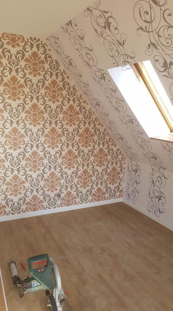 Pokój na poddaszu z wzorzystymi ścianami; jedna ściana w klasyczny wzór w odcieniach brązu, druga w delikatne, szare ornamenty; na podłodze leży ukośnica.