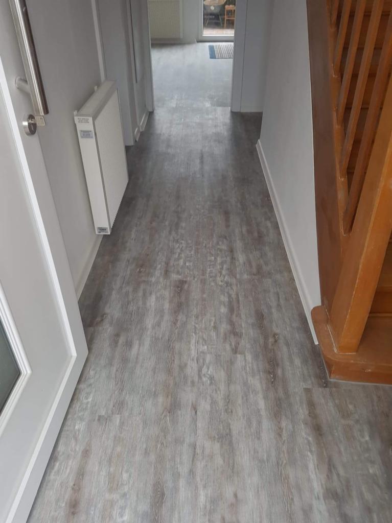 Ułożone panele podłogowe imitujące drewno w korytarzu, widoczny fragment schodów i grzejnik na ścianie.