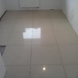 Jasne, błyszczące płytki podłogowe ułożone w kwadraty, z widocznym odbiciem okna i lampy, w narożniku widoczne rury instalacji centralnego ogrzewania.