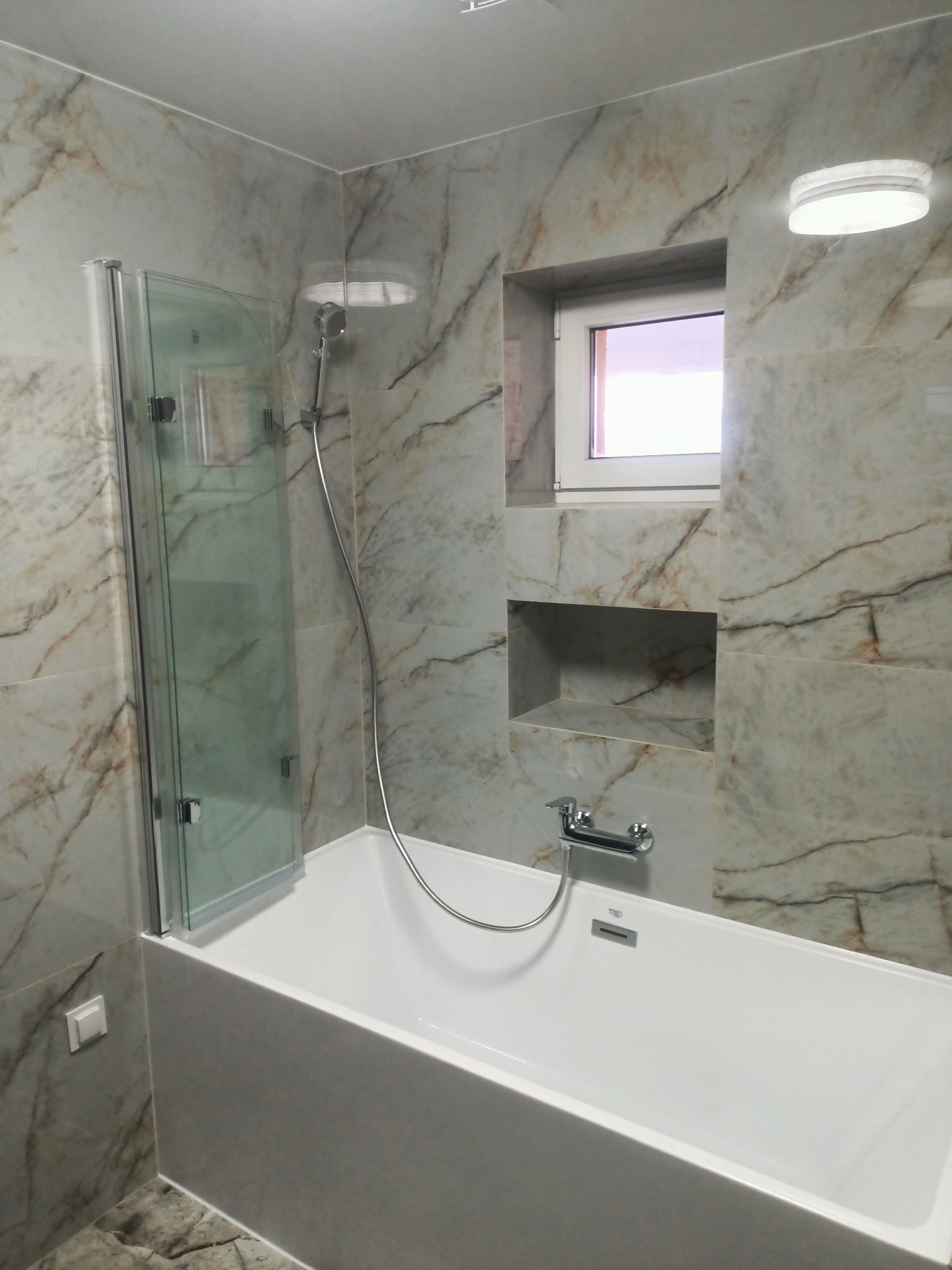 Nowoczesna łazienka z wanną, szklanym parawanem i marmurowymi płytkami. W ścianie wnęka z półkami i okno. Minimalistyczny design i jasne oświetlenie.