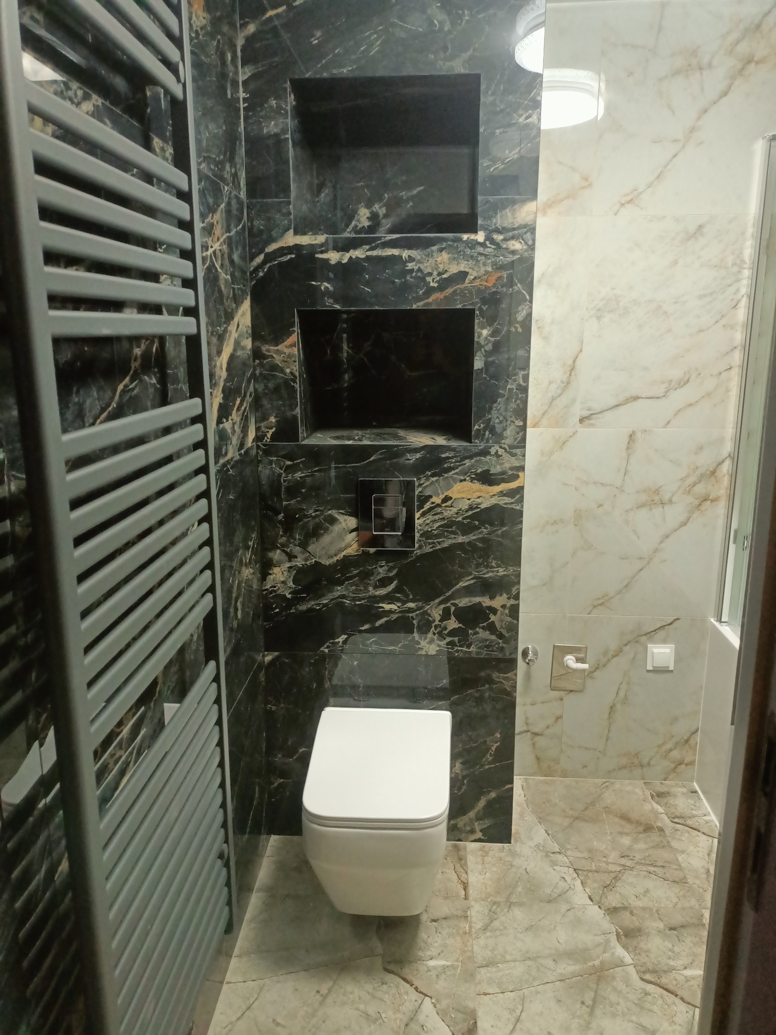 Wykończona łazienka w Jastkowie z marmurowymi płytkami, wiszącą toaletą i grzejnikiem drabinkowym. Ciemne wnęki dodają elegancji.