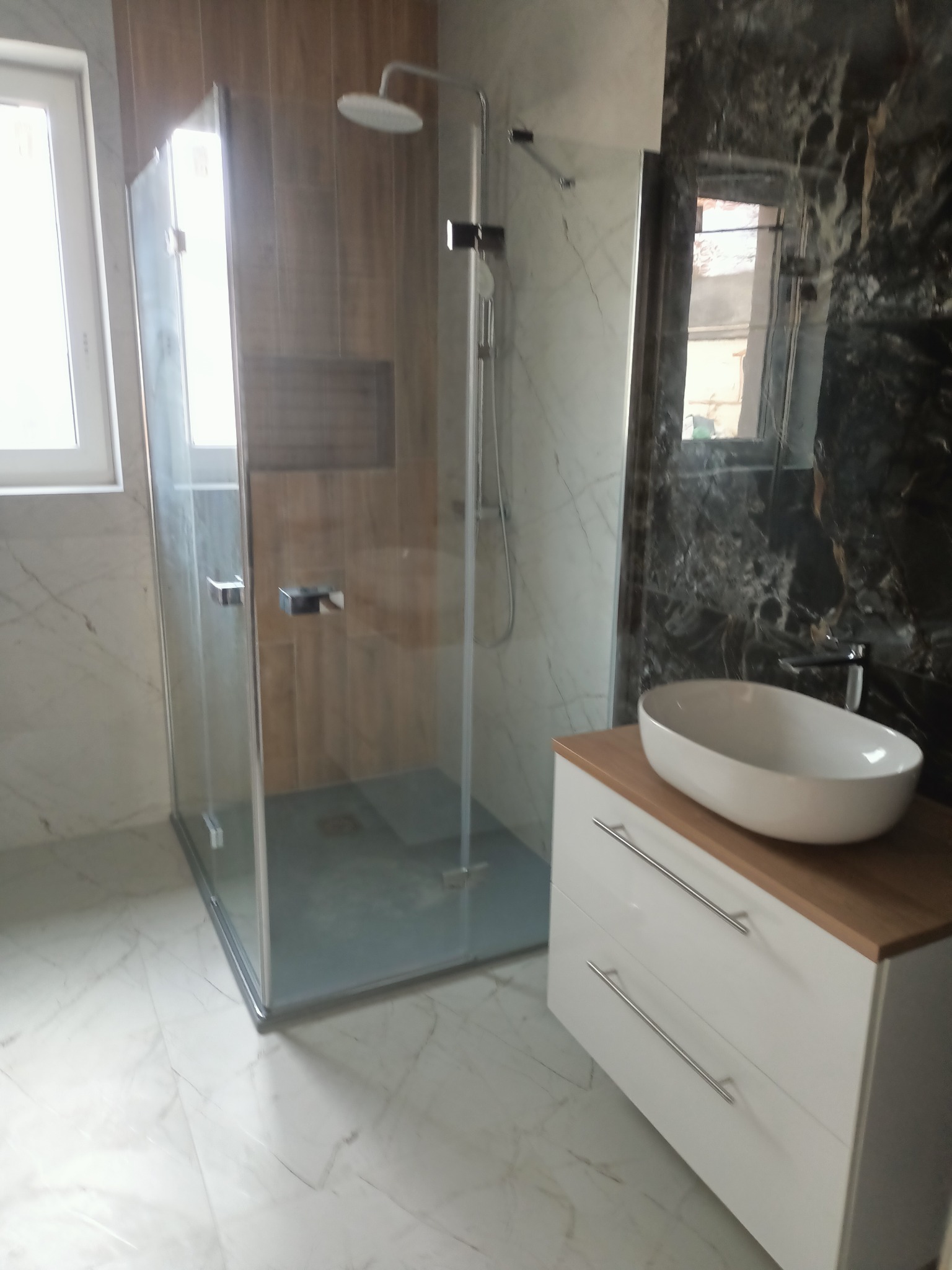 Nowoczesna łazienka z kabiną prysznicową typu walk-in, białą umywalką nablatową na szafce z drewnianym blatem i marmurowymi płytkami na ścianach.