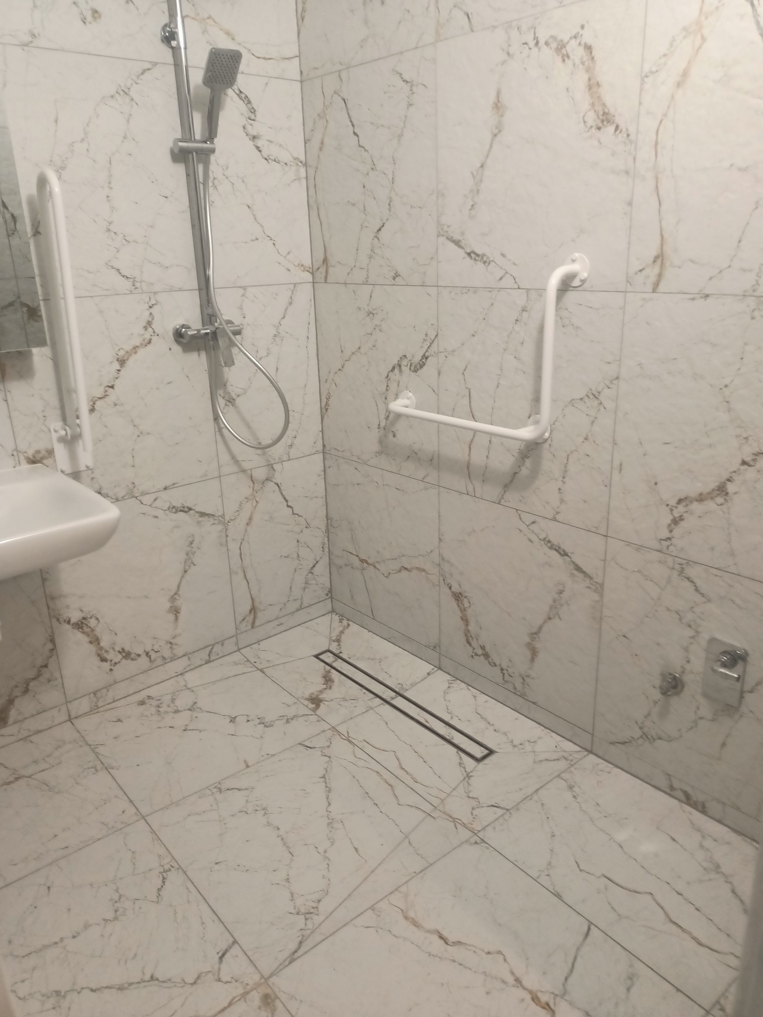Wykończona łazienka z marmurowymi płytkami, kabiną prysznicową, umywalką i uchwytami dla niepełnosprawnych. Nowoczesny odpływ liniowy w podłodze.