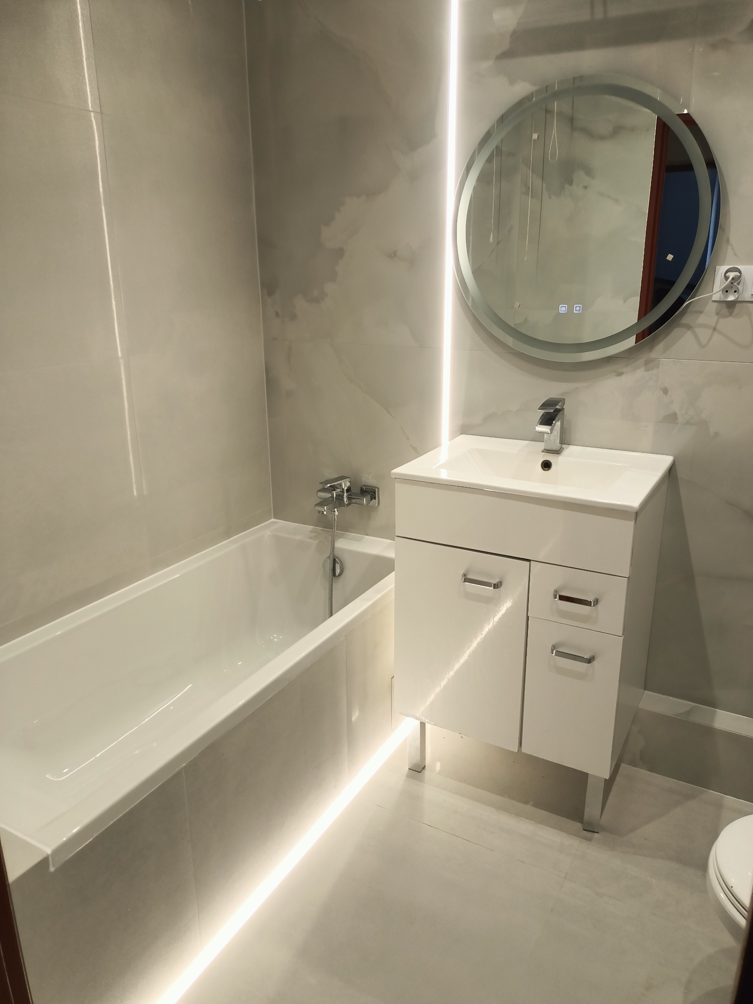 Nowoczesna łazienka z wanną, umywalką z szafką i okrągłym lustrem podświetlanym LED. Szare płytki imitujące marmur. Oświetlenie punktowe LED pod wanną.